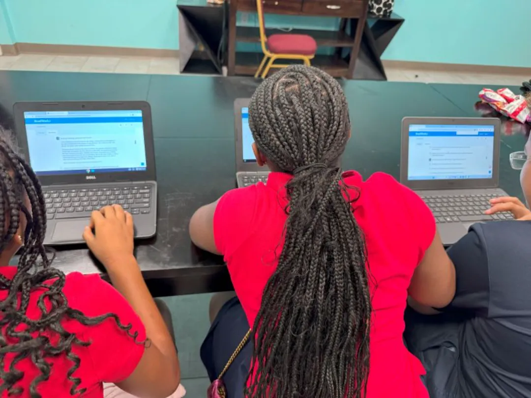 Rear view of three students with braided hair working on Chromebooks at a long black table, displaying reading comprehension or writing exercises.