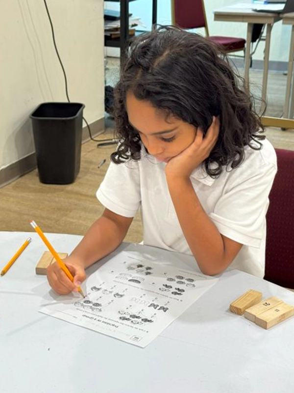 A young student with long dark hair sits at a table, resting her head on her hand while concentrating on a math or counting worksheet with a pencil.